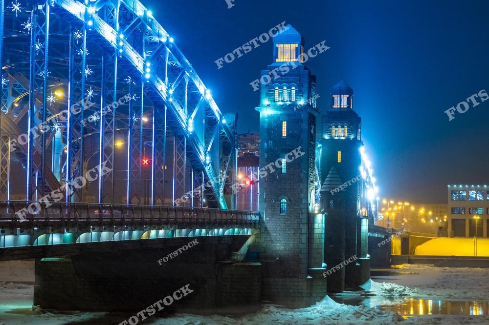 На фотографии изображен Большеохтинский мост в Санкт-Петербурге, ярко подсвеченный ночными огнями. Сложные металлические конструкции моста украшены световыми элементами, создавая эффект сказочной иллюминации. Ледяная поверхность Невы усиливает ощущение зимней прохлады. Этот мост – один из символов инженерного искусства и красоты ночного Петербурга.